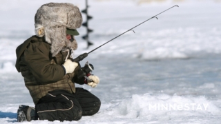 Child ice-fishing in Minnesota seen with a fishing pole in a hole, and a big fuzzy hat.