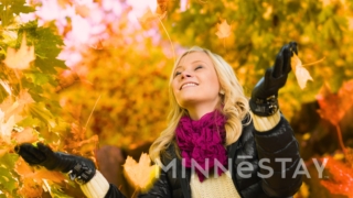 Woman throwing colorful fall leaves in the air with beautiful fall foliage in all its glory behind her.
