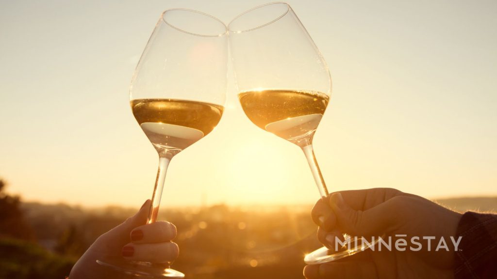 Romantic Escape with MINNeSTAY two glasses of white wine clinking together at sunset.